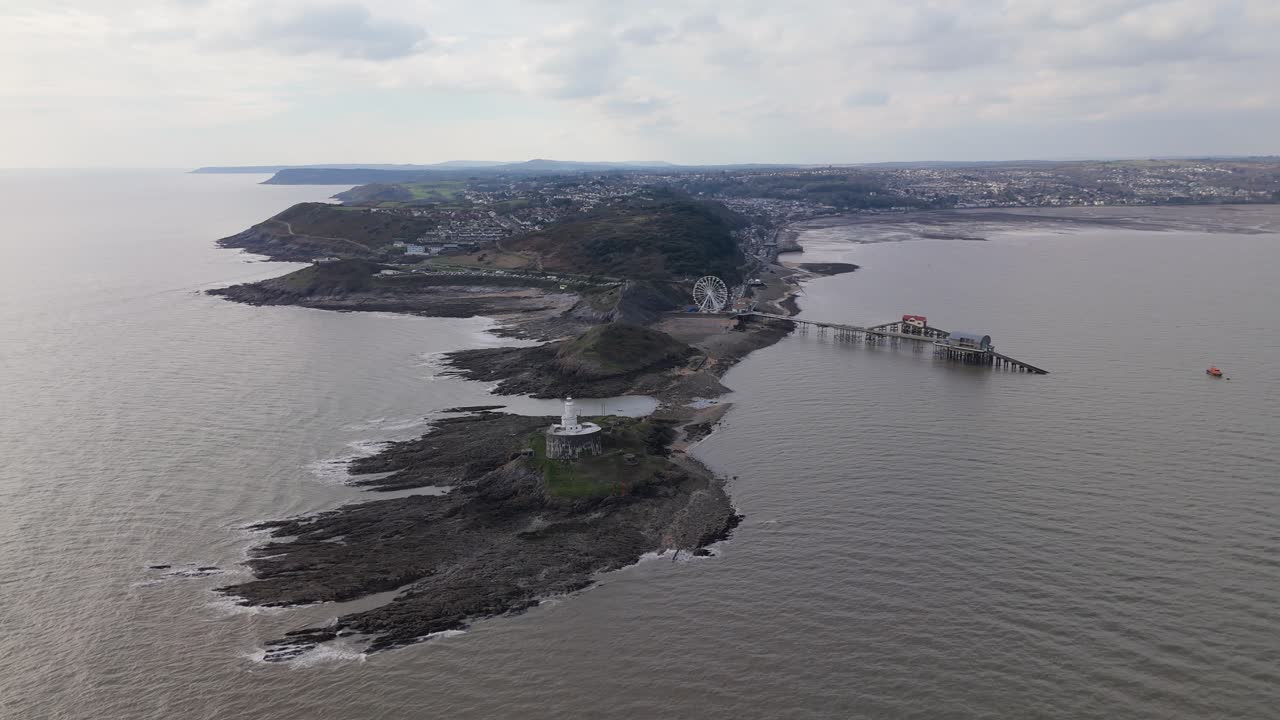A moody aerial orbit counterclockwise of Mumbles Pier, with the Big Wheel overlooking the coastline under a cloudy sky