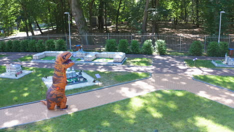 orange tirex dinosaur having a walk nearby miniature buildings. dinosaur turning around himself and walking away. nature park backdrop.