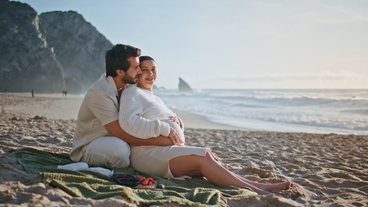 임신 한 커플 이 해변 에 앉아 여름 피크닉 을 즐기고 있다. 남편 이 아내 의 배 를 포옹 하고 있다