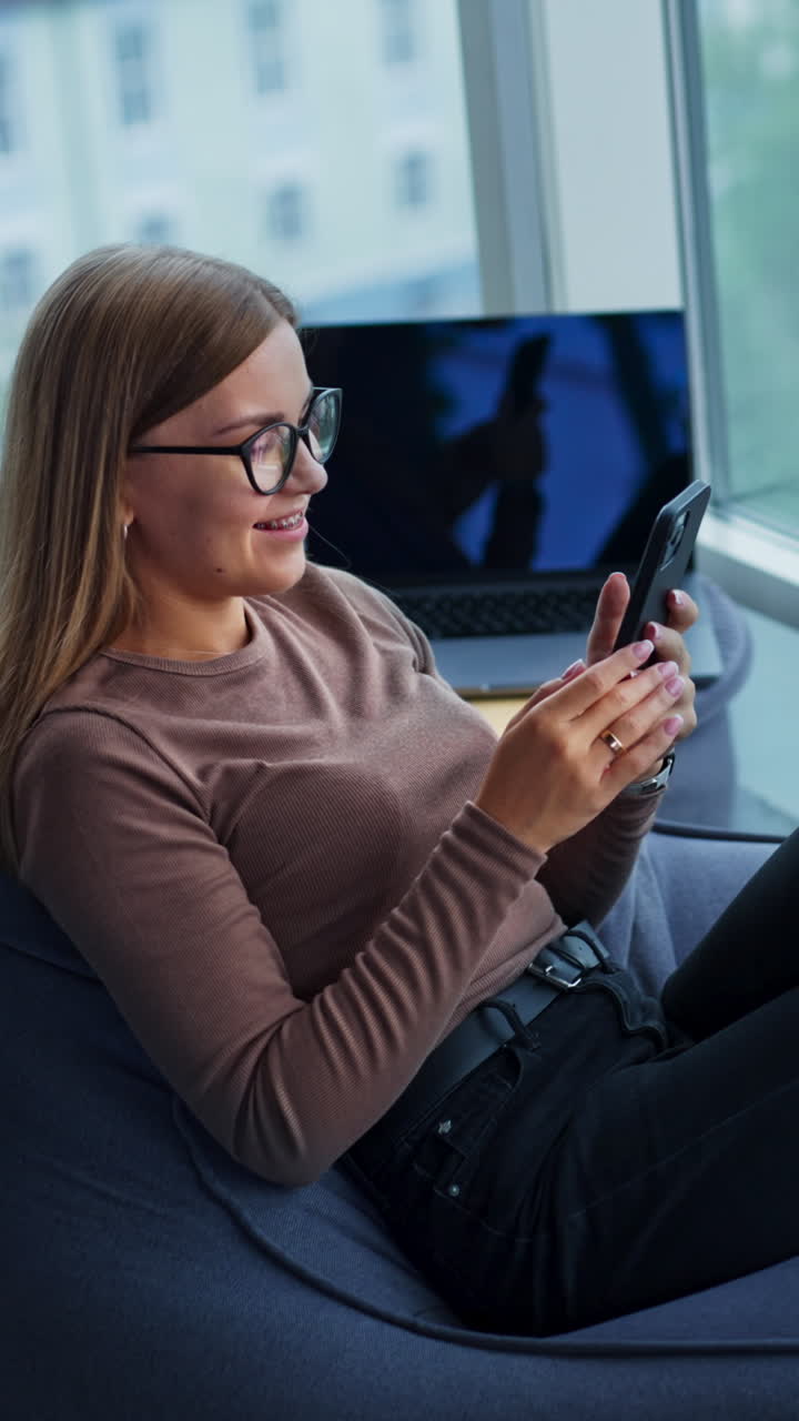 Relaxed long-haired lady wearing glasses sits in bean bag chair. Woman looking at her phone and then taking selfie. Blurred cityscape at backdrop. Vertical video