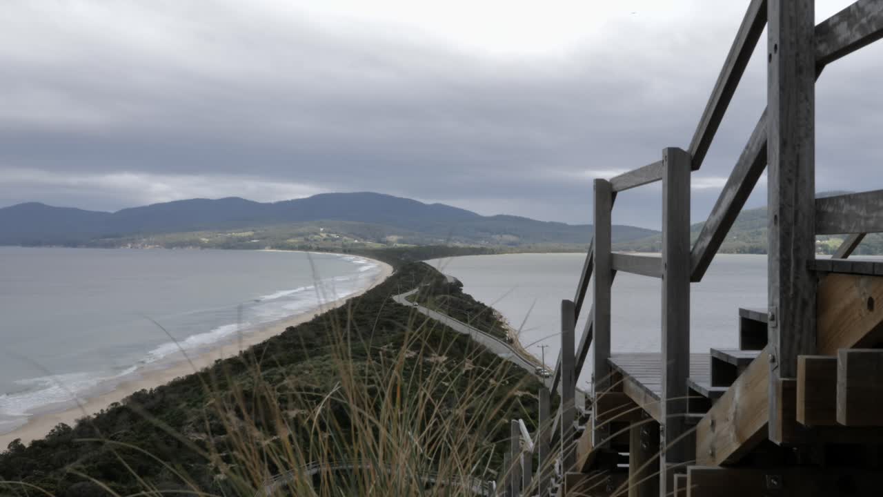 el viento sopla arbustos con vistas al istmo de long island rodeado de olas de playa oceánica y montañas verdes y bosques en un día nublado nublado mientras el automóvil conduce por la carretera, isla bruny, tasmania, australia