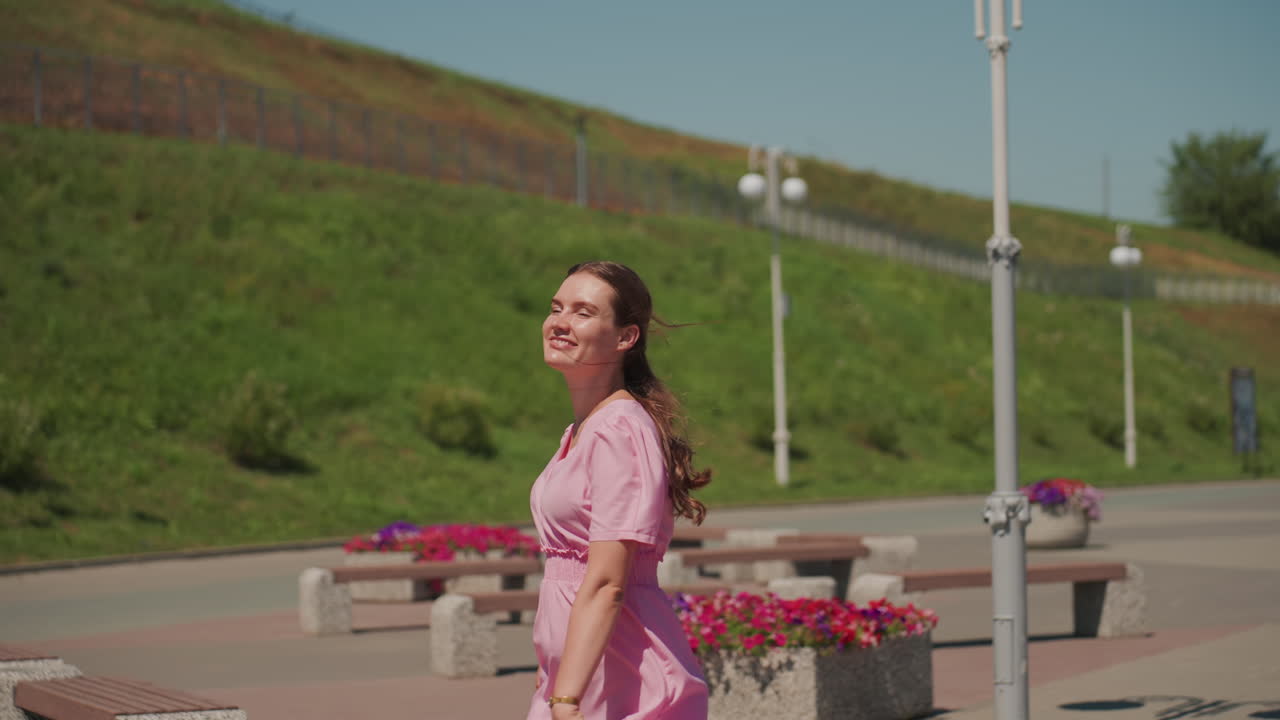 Una joven con un vestido rosa pasea por un paseo marítimo iluminado por el sol, su cabello trenzado ondea con la brisa, con una sonrisa mira hacia arriba, sus ligeros pasos dejan atrás farolas y jardineras, un terraplén verde y un entorno urbano moderno
