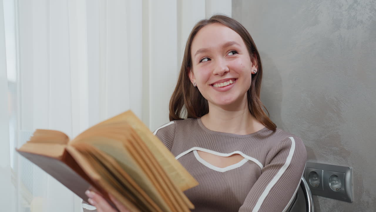 joven sentada en la mesa leyendo un libro mira hacia arriba con una cálida sonrisa, creando una atmósfera pacífica y acogedora en un interior bien iluminado con cortinas suaves en el fondo