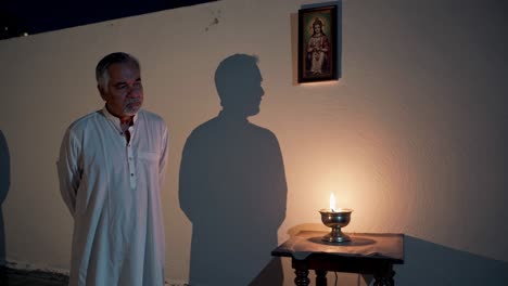 elderly man standing with hands behind his back, bathed in the warm glow of an oil lamp, engaging in prayer or contemplation in a dimly lit room