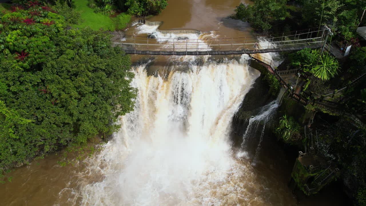 vista aérea de mena creek fall y una mujer caminando por un puente peatonal sobre el agua en un caluroso día soleado, australia