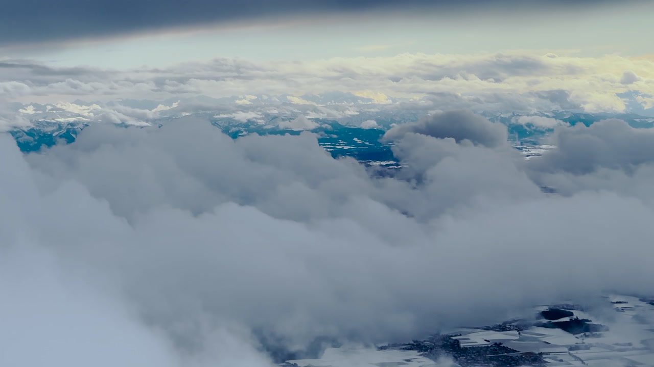 Airplane window view of snow-covered Bavarian Alps with dramatic winter clouds and frozen landscape, Germany