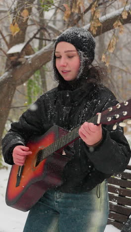 young woman in black hoodie playing guitar outdoors in snowy park, standing near snow-covered bench, surrounded by frosty trees and softly falling snow