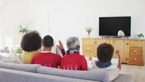 Biracial-family-watching-tv-with-rugby-ball-at-stadium-on-screen