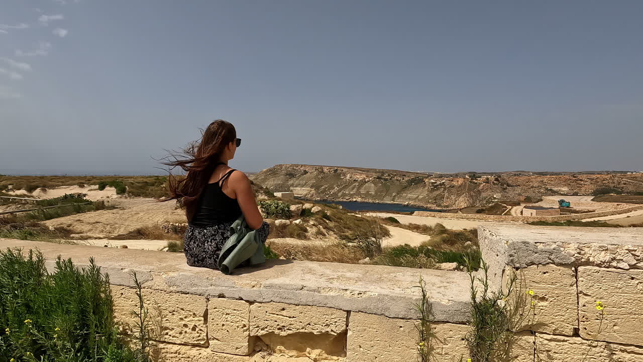 mujer sentada en una pared de piedra con un paisaje desértico detrás y el viento soplando su cabello