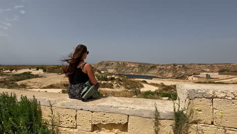 woman sitting on a stone wall with a desert landscape behind and the wind blowing her hair