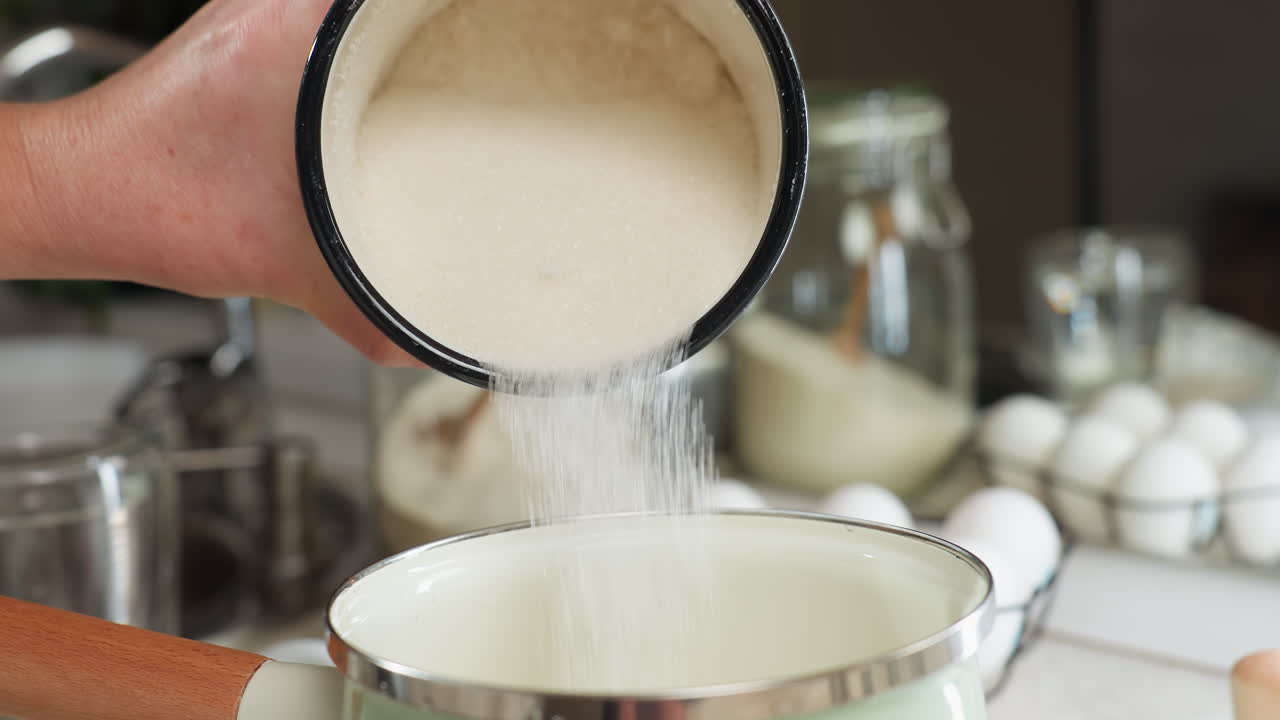 Close up of person pouring sugar from enamel cup into ceramic pot with wooden handle while egg rack, glass jars, and kitchen utensils surround cooking space