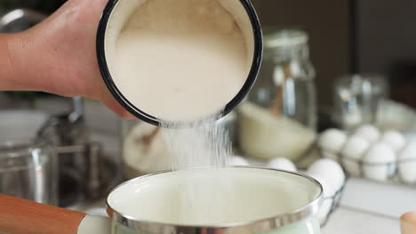close up of person pouring sugar from enamel cup into ceramic pot with wooden handle while egg rack, glass jars, and kitchen utensils surround cooking space