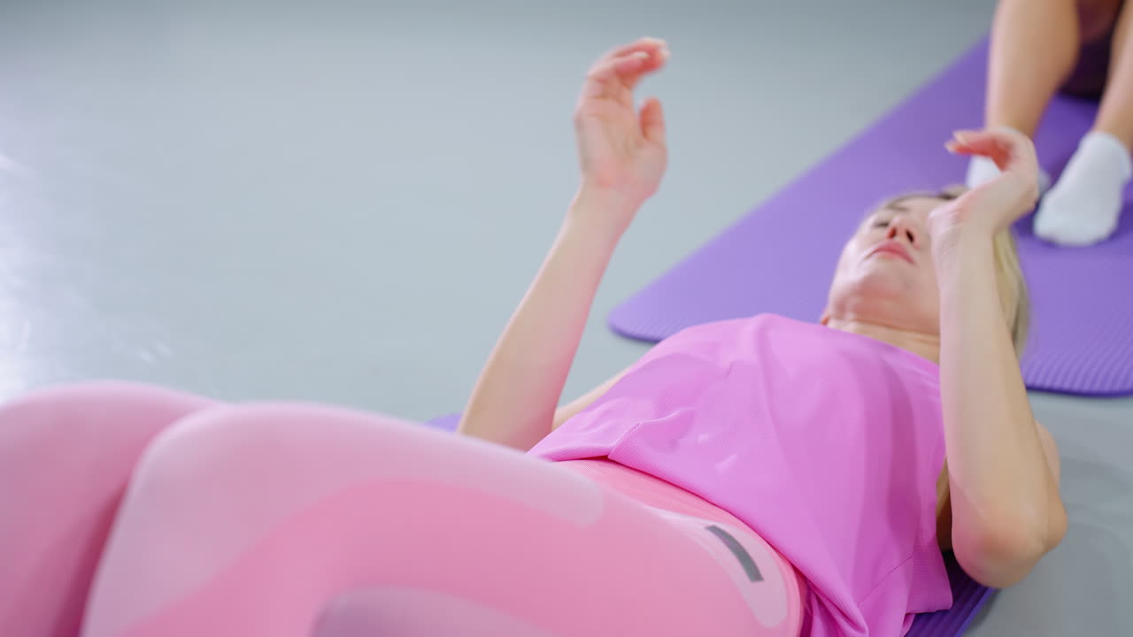 женщина в розовом тренировочном костюме выполняет сидячие упражнения, откидываясь назад и протягивая руки вперед, сосредоточенное выражение лица показывает решимость, другой человек на заднем плане