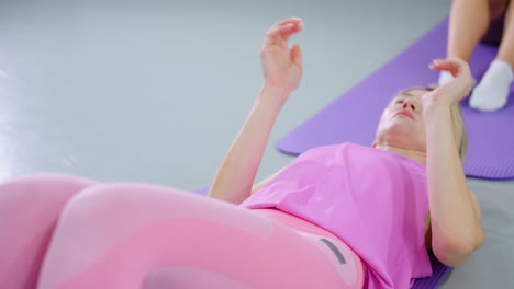 mujer con traje de entrenamiento rosa realiza un ejercicio de sentada, inclinándose hacia atrás y estirando los brazos hacia adelante, la expresión enfocada muestra determinación, otra persona en el fondo