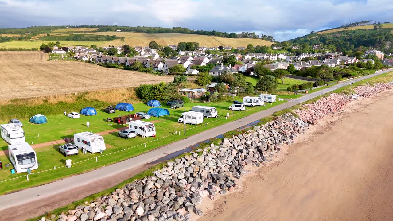 Drone footage glides above a seaside campsite with caravans, tents, and motorhomes along a rocky shoreline in Rosemarkie, Highland, Scotland. Bright daylight, wide shots