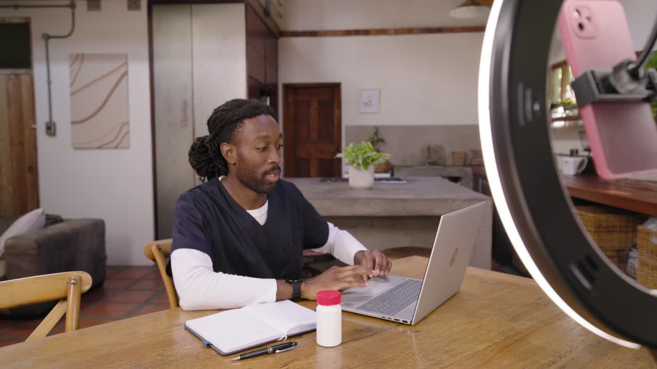 Erwachsener afrikanischer Mann in Arbeitskleidung antwortet auf ein rosa Telefon mit Ringlicht, zeigt eine weiße Flasche und tippt am Laptop.
