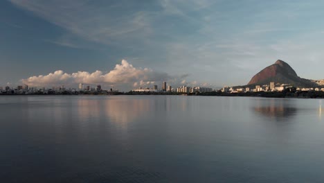 Aerial-upwards-movement-revealing-the-Rio-de-Janeiro-city-lake-at-blue-hour-mirroring-a-statue-on-a-rock-and-going-beyond