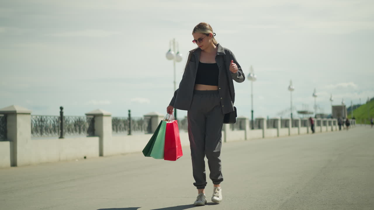dama con camiseta negra bajo chaqueta gris baila alegremente, sosteniendo bolsas de compras rojas, menta y verdes en su mano derecha, su sombra es visible en el suelo, con personas caminando en el fondo