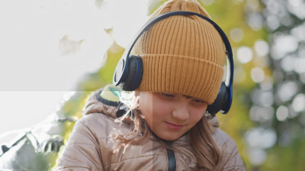 close up of girl wearing mustard beanie and black headphones sitting on ornate metal bench in autumn park gently manipulating yellow leaf in lap as soft bokeh foliage