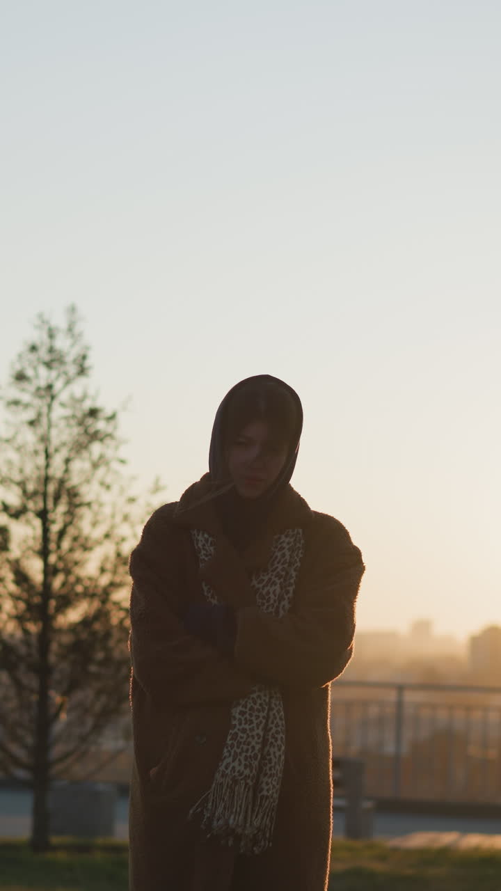 una niña afligida está de pie frente a un monumento de piedra en un parque durante la puesta del sol, sus manos cruzadas en sus brazos, con un abrigo y una bufanda. la cálida luz dorada realza el estado de ánimo triste