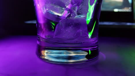 Ice-cubes-and-drink-poured-into-a-glass,-under-blue-and-purple-lights,-macro-shot,-close-up-view