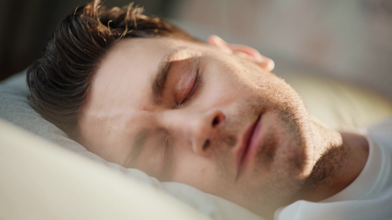 Giovane uomo che dorme sul cuscino alla calda luce del sole. Primo piano del viso, ritratto di riposo sereno.