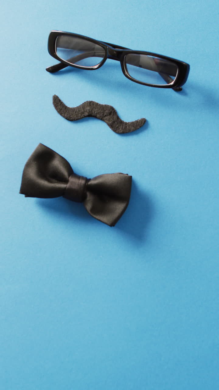 Vertical video of black false moustache, glasses and bow tie on blue background with copy space