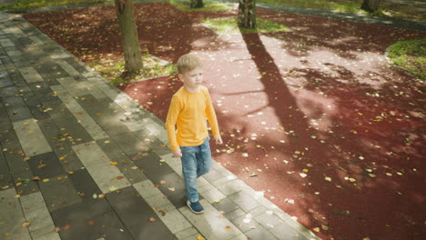 young boy in orange long-sleeve shirt and jeans walking confidently along paved path in leafy park, surrounded by trees and scattered autumn leaves under sunlight casting playful shadows