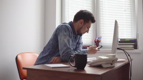 Joven-Oficinista-Usando-Su-Teléfono-En-La-Oficina-Sentado-En-La-Mesa-Con-Computadora,-Teléfono-Y-Taza.-Filmado-En-4k