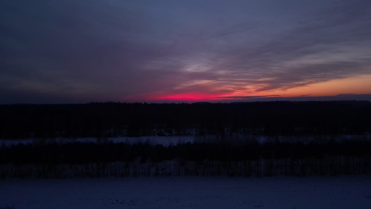 4K-luftoptagelse, der trækker sig væk fra en mørk skovvæg i den dybe skumring. Lyserødt tusmørkebånd hænger over horisonten med en kølig, blå øvre himmel og svage, varme toner til højre over vintermarkerne