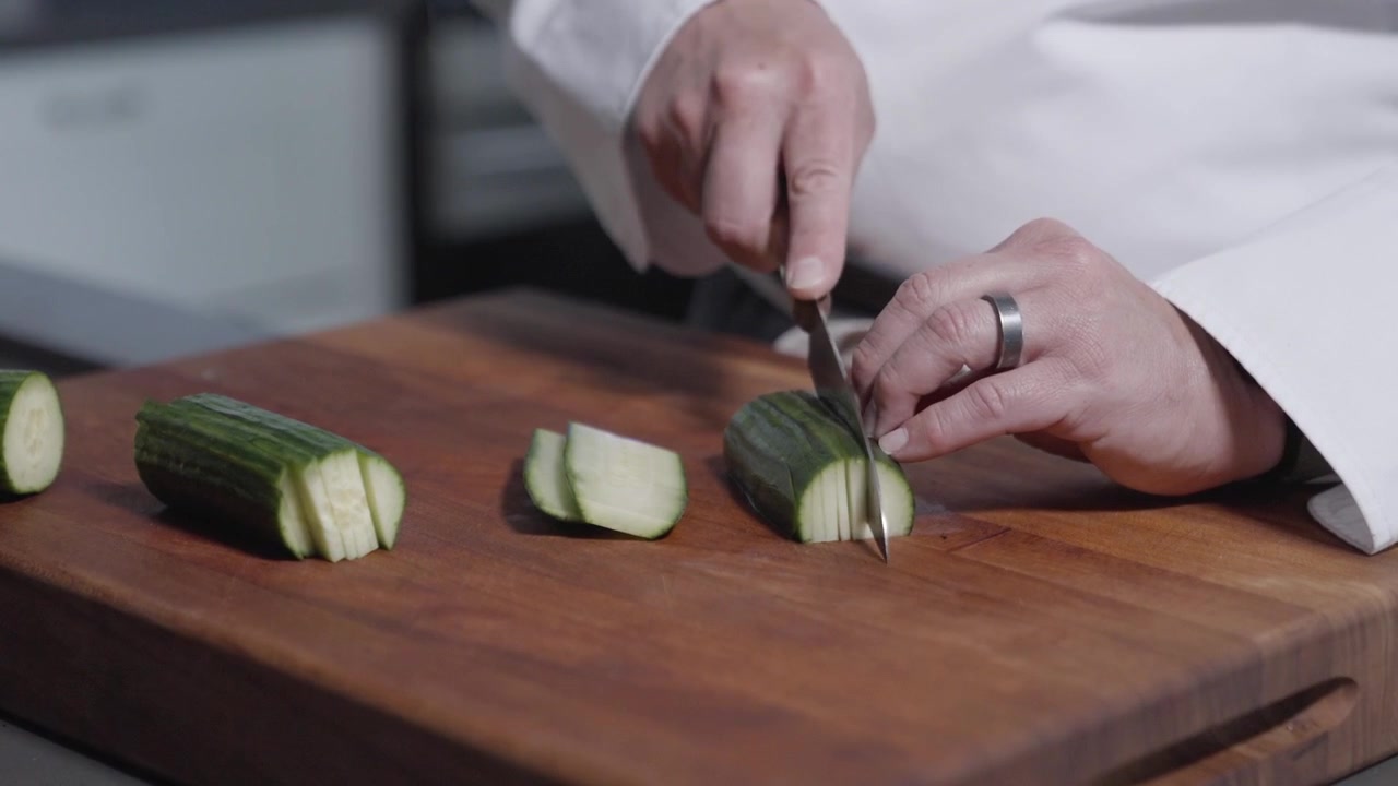 Una ripresa statica di uno chef che affetta con maestria più cetrioli per il lungo su un tagliere. Perfetto per contenuti culinari, evidenziando le abilità con il coltello e le tecniche di preparazione del cibo in una cucina professionale