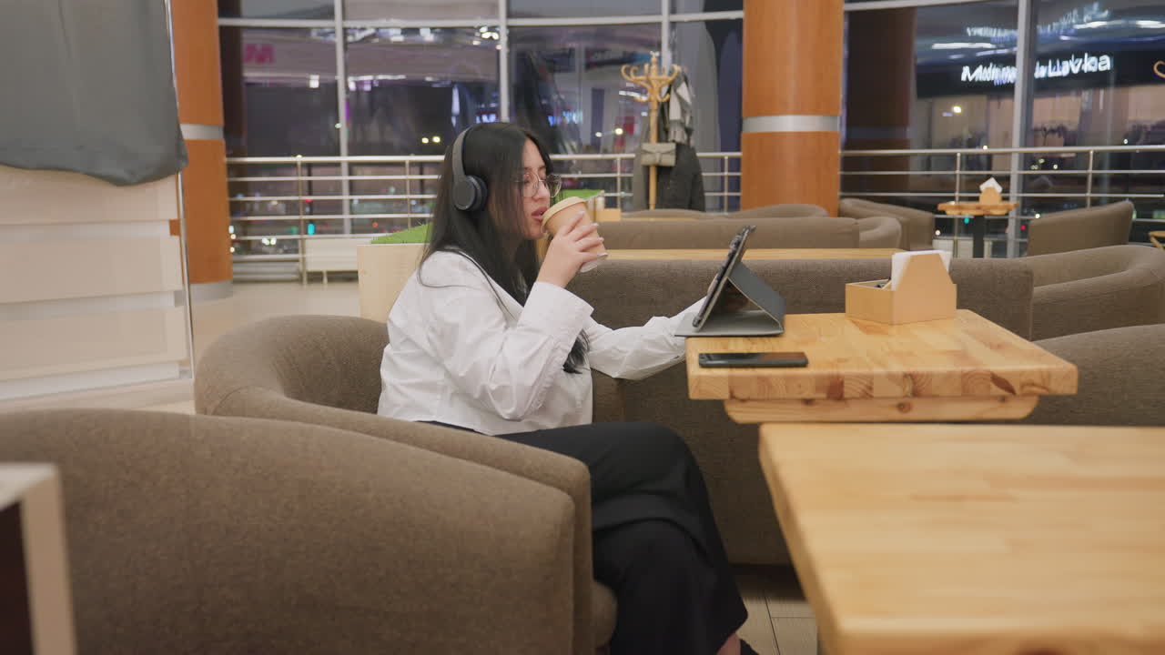 lady wearing headphones sits in cafe sipping coffee while holding tablet in one hand, surrounded by modern interior with warm lighting and reflections from evening city view through large windows