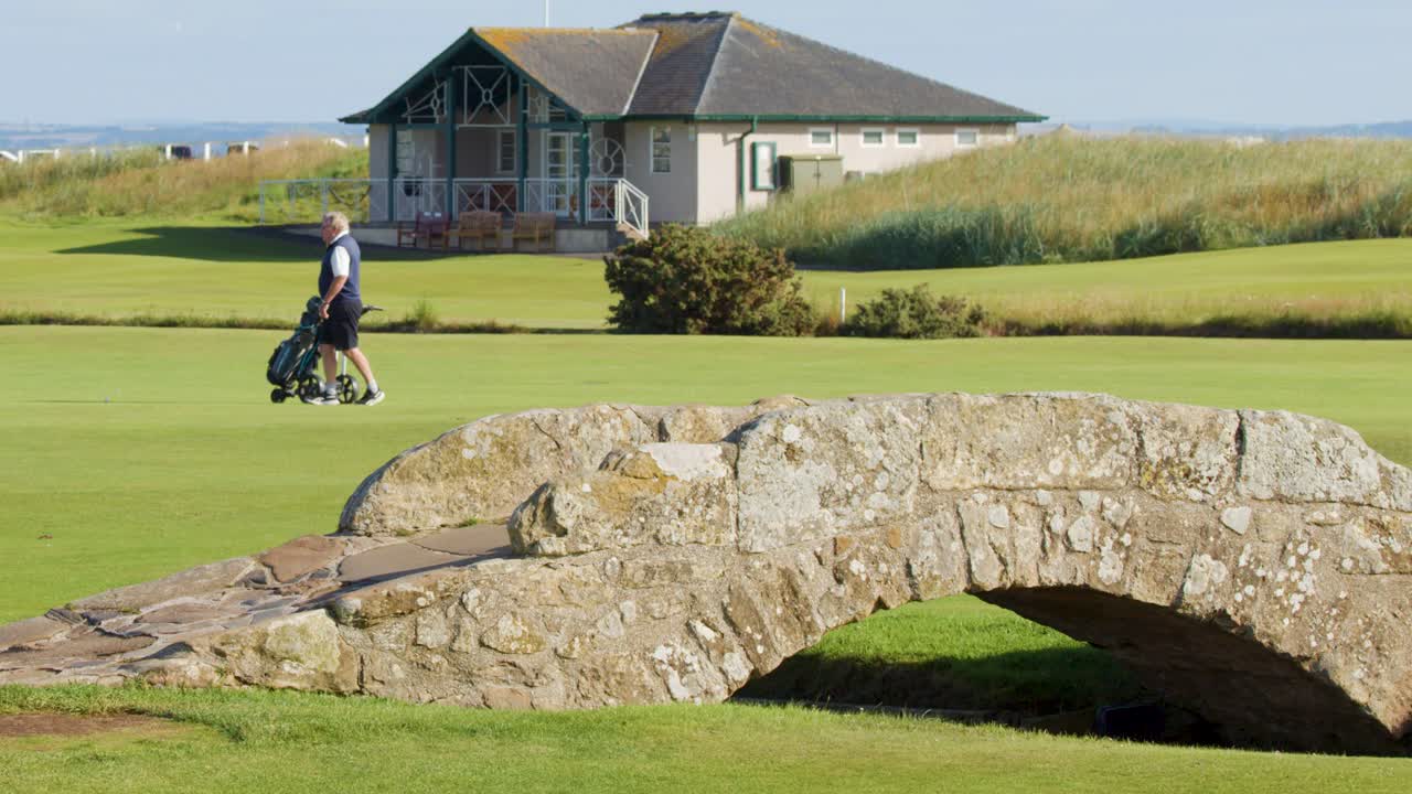 An elderly man pushes a golf trolley across a historic stone bridge on a lush golf course in bright daylight at St Andrews, Scotland