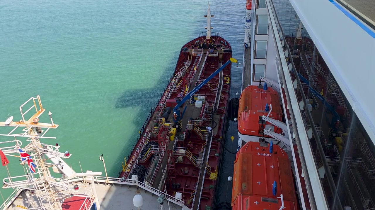 Carnival Legend alongside a bunker tanker during a marine refueling operation in Dover, England, with tanker deck piping, loading equipment and orange lifeboats visible from the cruise ship