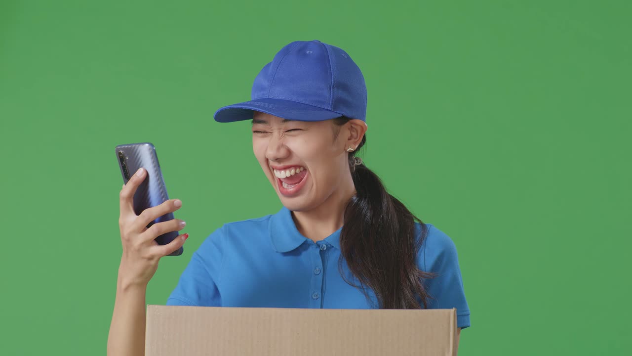 Close Up Of Asian Female Courier In Blue Uniform Looking At Smartphone And Celebrating The Succeed While Delivering A Carton On Green Screen Background In The Studio