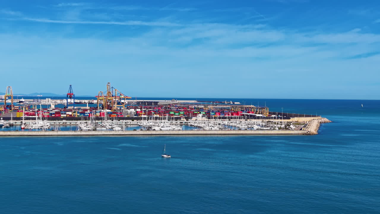 Container port in Valencia, Spain, with stacked shipping containers, cranes, and a marina filled with sailboats along the Mediterranean coast under blue skies