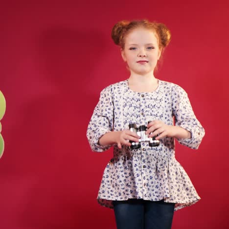 little girl in print blouse are looking through binoculars around herself on the background of red studio and scenery of trees