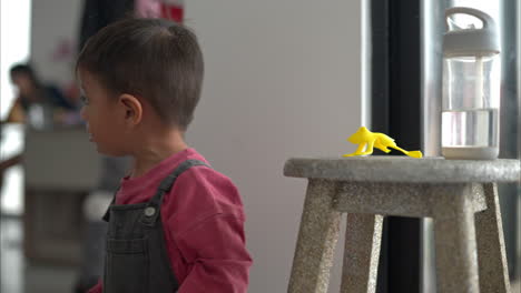 little latin hispanic boy playing with a yellow broken plastic frog toy on a wooden stool sneezing due to dust in the air