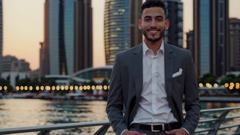 confident young businessman wearing tailored suit standing near river, smiling with modern skyscrapers and sunset cityscape behind, embodying professional success and urban lifestyle