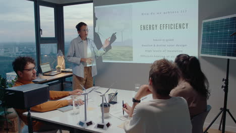 grupo de colegas escuchando la presentación de eficiencia energética dada por una ingeniera en una reunión de negocios en una oficina con ventanas panorámicas con vistas a la ciudad