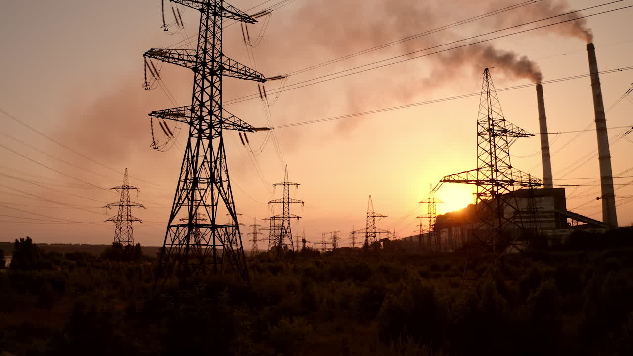 Electric towers near factory chimney with dark smoke. Transmission lines on background of harmful fumes from industry releasing from pipes at sunset. Environment pollution