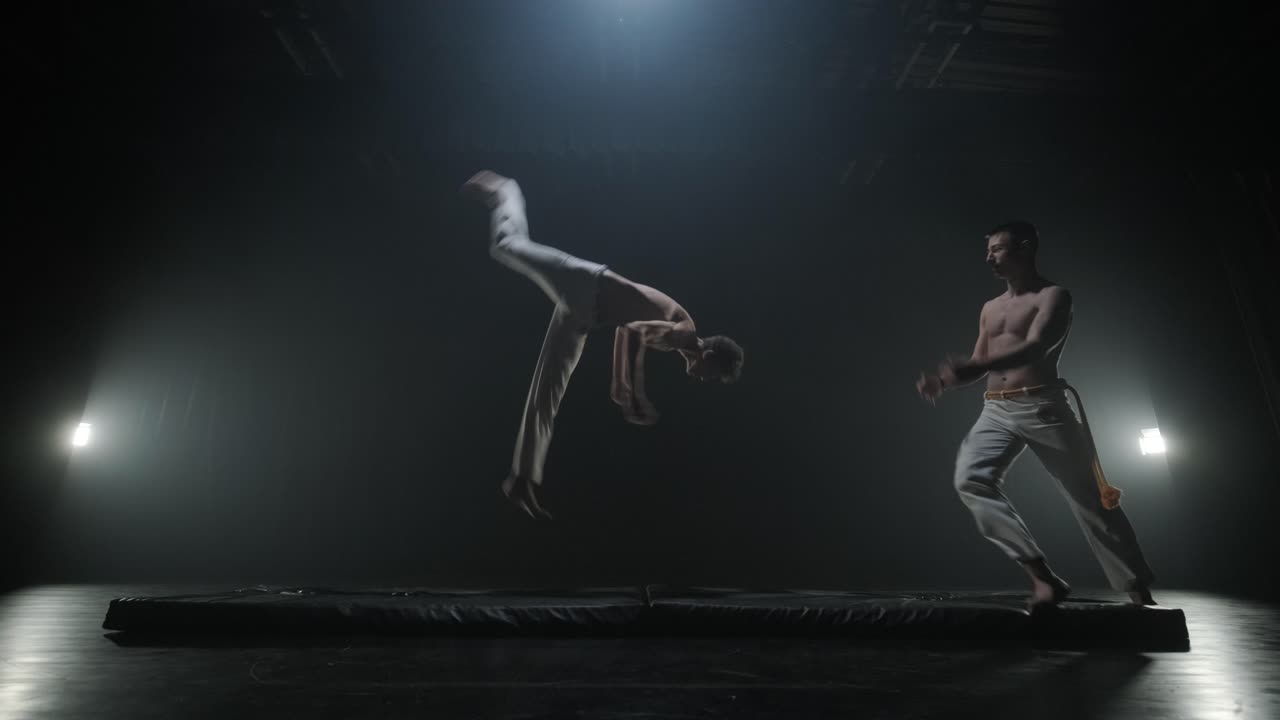 un grupo de bailarines profesionales están practicando capoeira en la oscuridad contra un foco de luz en un fondo negro de estudio. arte marcial afro-brasileño que combina elementos de la danza.