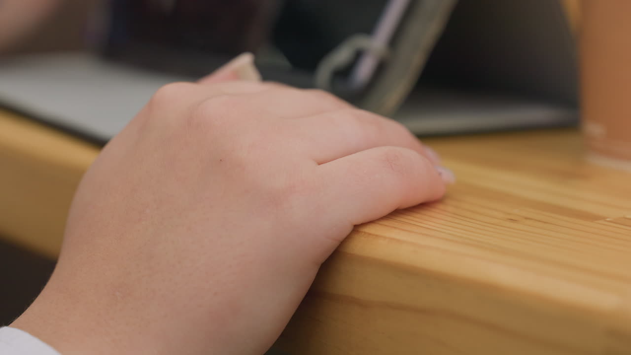close up of young woman fingers gently tapping on wooden table in calm setting with soft lighting and modern indoor background creating focused yet relaxed atmosphere near tablet and coffee cup