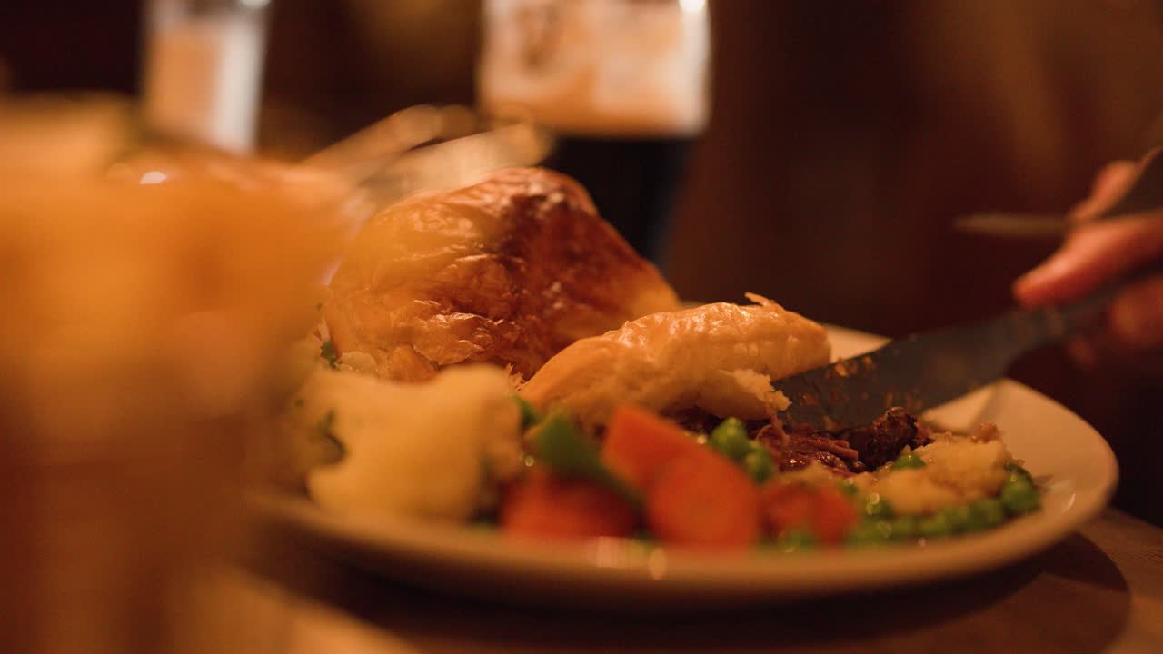 Hand slices steak pie with vegetables under warm lighting, shallow depth of field, intimate setting