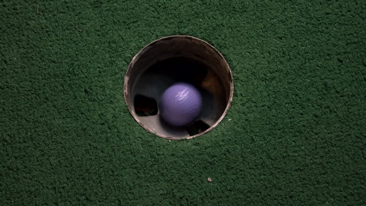 A close up birds eye view of a purple mini golf ball falls into the golf hole and bounces in the pocket on a course