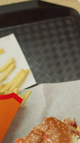 primer plano de papas fritas, hamburguesa y taza de café en una bandeja negra en una mesa de madera marrón, mientras una mujer con un vestido rosado deja caer la bebida de nuevo en la bandeja sostiene una fritura a mitad de comer en la mano