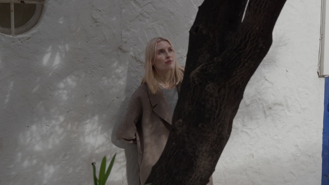 Blonde Caucasian woman leaning against a shadowy wall, looking through the leaves and both directions
