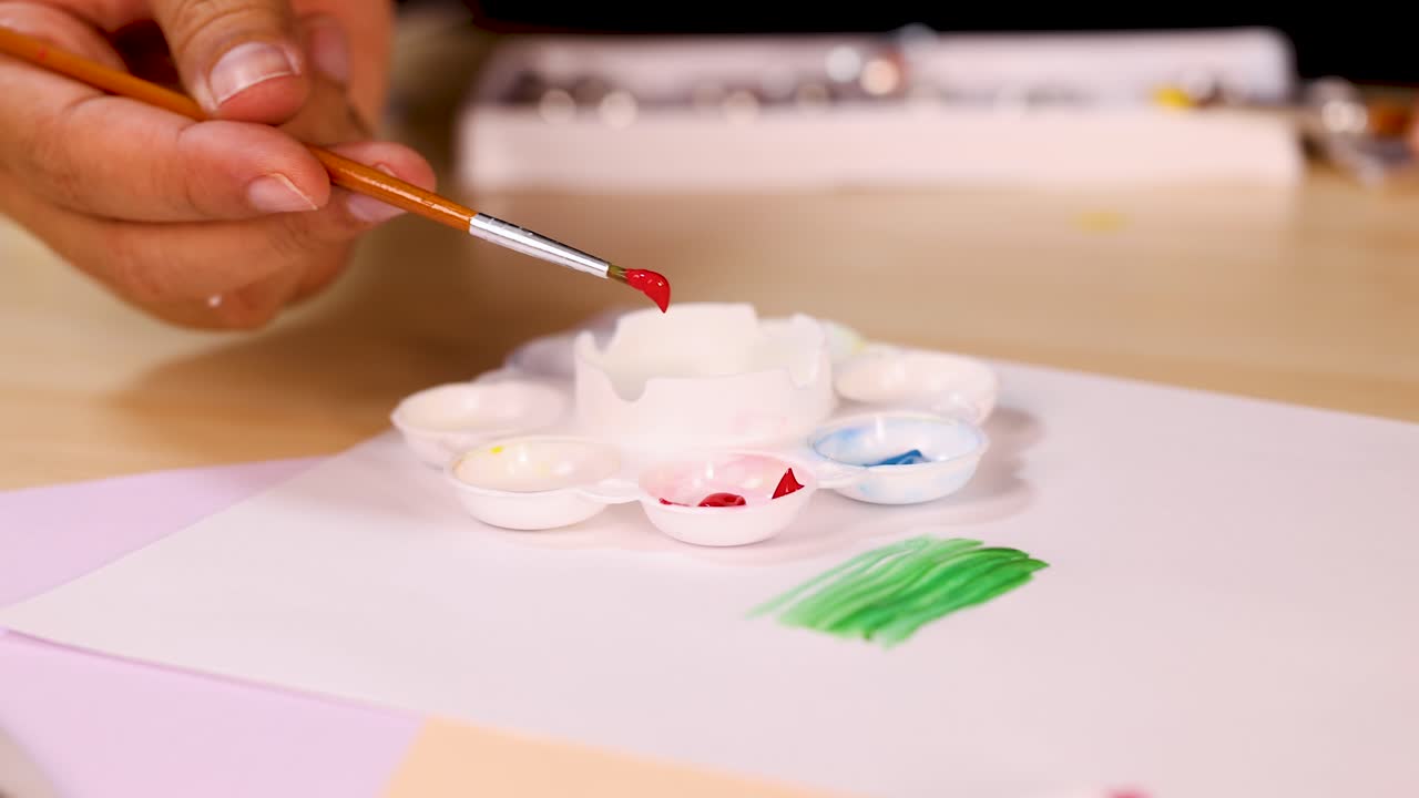 Hand mixes red watercolor paint on palette with brush, close-up, warm indoor lighting, stationary camera