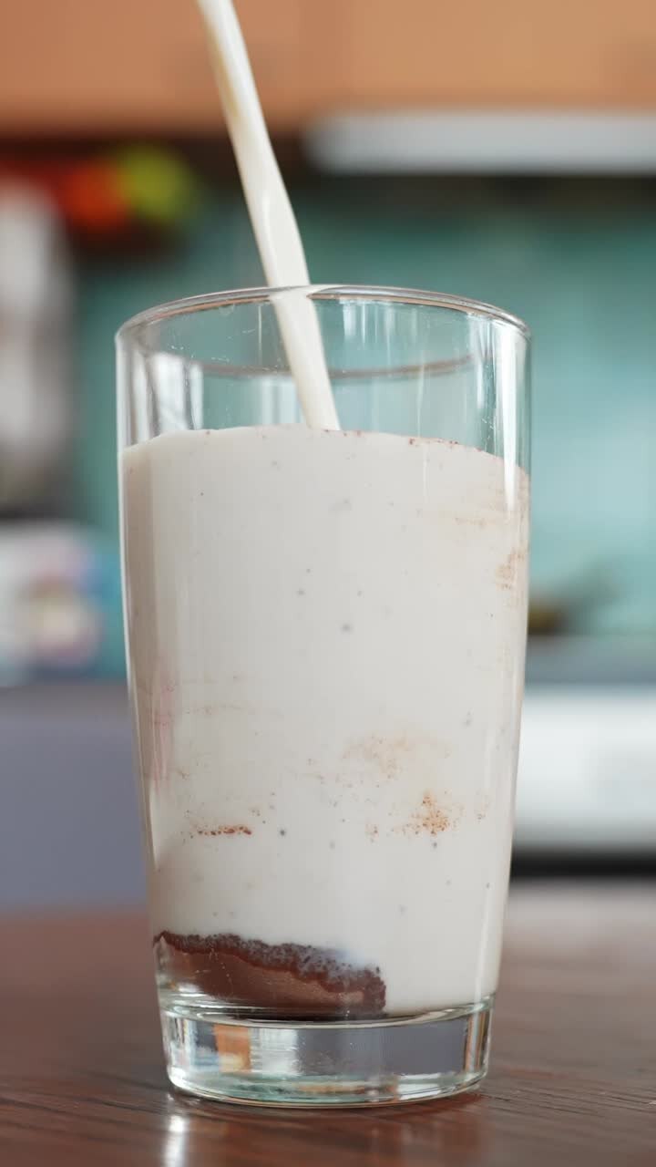 Primo piano verticale di latte versato in un bicchiere alto con cacao in polvere, preparazione di latte e cioccolato fatto in casa su un tavolo di legno con sfondo di cucina tenue e spazio per il testo