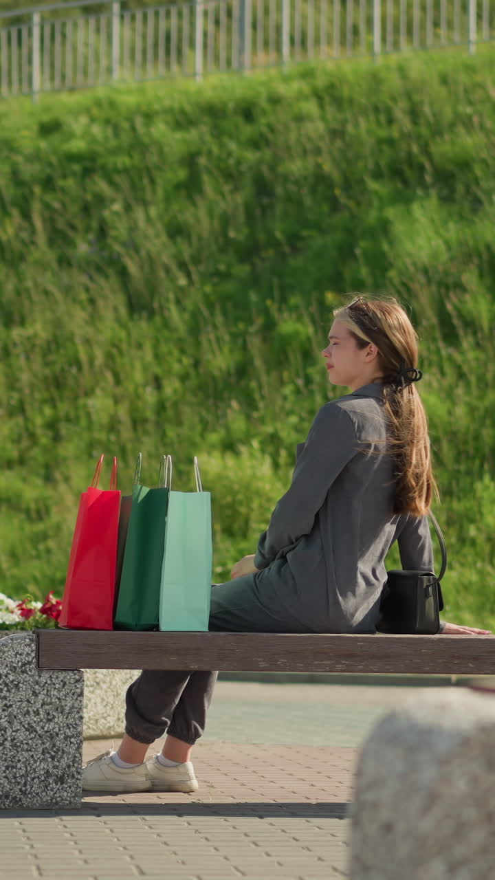 여자는 벤치에 걸어, 다채로운 쇼핑 가방을 떨어뜨리고, 좌석에 앉아, 왼손을 편안하게 하고, 오른쪽 다리를 앞으로 움직여서 멀리 바라보고, 언덕의 초록색이 배경에 보인다.